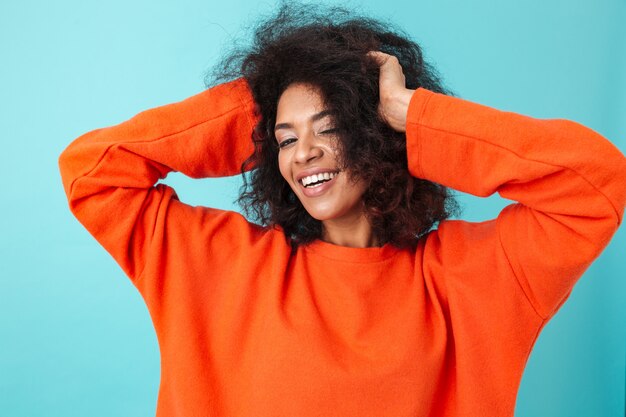 Colorful portrait of smiling woman in red shirt posing  and touching her dark curly hair, isolated over blue wall