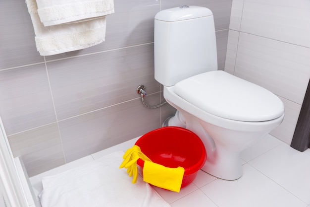 Colorful red basin, yellow gloves and cloth for cleaning in a bathroom standing on the floor alongside a plain white toilet