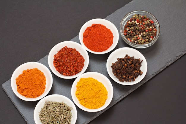 Colorful various ground spices, dry cloves and herbs in bowls on black stone cutting board. Top view.