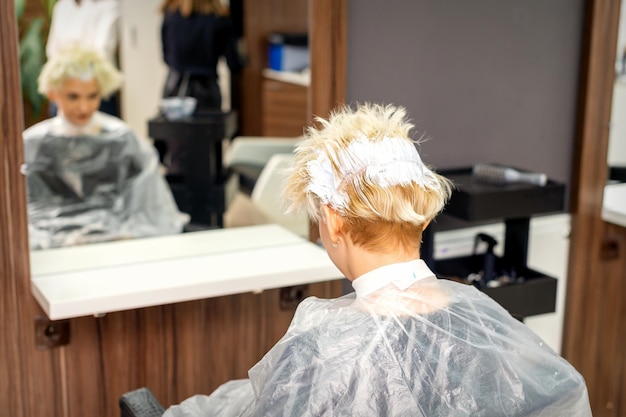 Coloring white hair with hair dye of the young caucasian blonde woman sitting at a hair salon close up