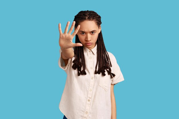 Do not come closer. Serious annoyed woman with black dreadlocks keeps palm in stop gesture, asks not to bother her, looks angrily, wearing white shirt. Indoor studio shot isolated on blue background.