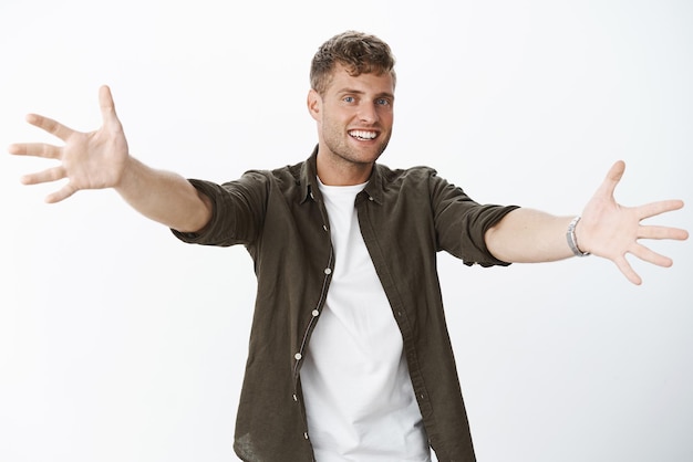 Come here hug me Portrait of friendly and joyful charismatic european guy with blue eyes and blond stylish haircut extending arms sideways to cuddle reaching at camera with broad smile over grey wall