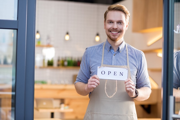 Come to our cafe. Happy delighted friendly man smiling to you and holding the label tag while inviting customers