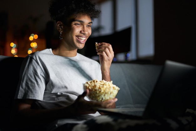 Comedy happy young guy smiling watching movie online using laptop and eating popcorn while sitting