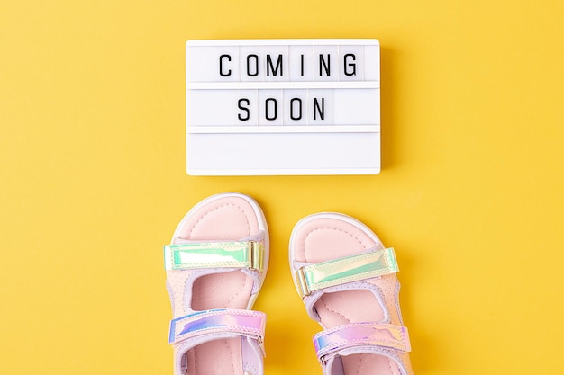 Coming soon Motivational quote on lightbox and stylish holographic sandals on yellow background Top view Flat lay Creative inspirational summer concept