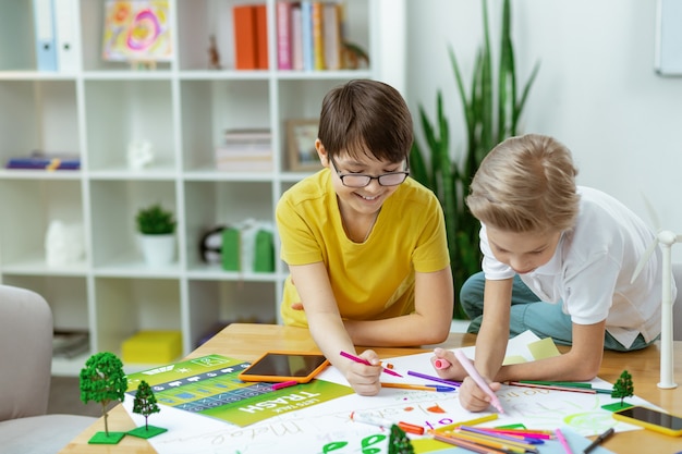 Communicative young boys. Cheerful dark-haired boy in yellow t-shirt using colorful markers while entertaining himself with drawing