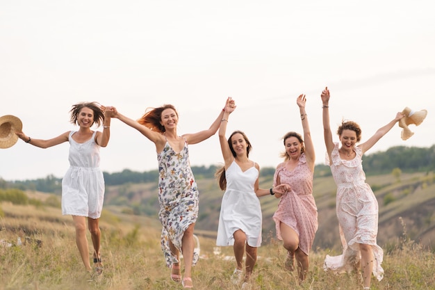 The company of cheerful female friends have a great time together on picnic in a picturesque place overlooking the green hills. Girls in white dresses dancing in the field