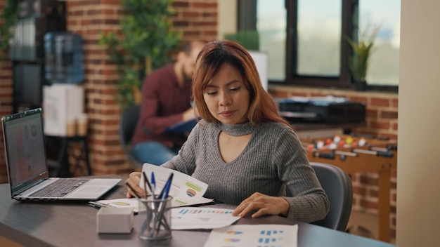 Company employee analyzing documents to plan marketing strategy in startup office. Business woman looking at financial charts on papers and laptop, developing sales investment.