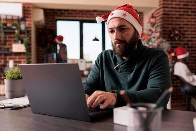 Company employee with santa hat working on laptop in festive office, christmas tree lights and decorations. Using computer in workplace with seasonal holiday ornaments and xmas decor.