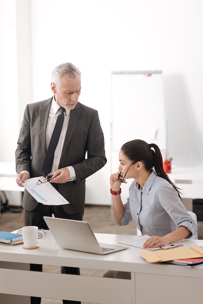 Competent young woman holding glasses in right hand, putting her left hand on the table while looking at documents