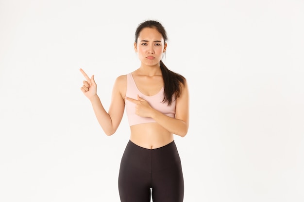 Complaining sad asian girl in activewear, sulking and frowning upset as pointing upper left corner, standing white background.