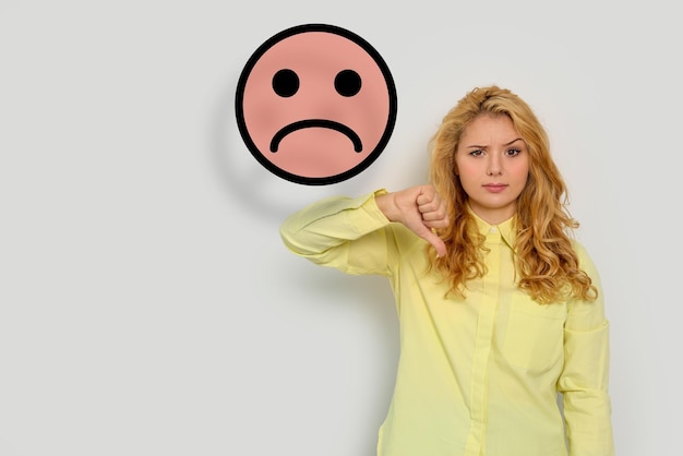 Photo complaint dissatisfied woman showing thumbsdown on white background illustration of sad face near her