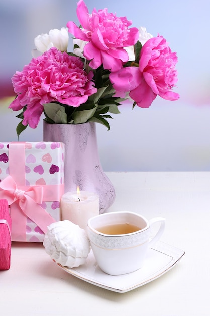 Composition of beautiful peonies in vase, tea in cup and marshmallow, on table, on light background