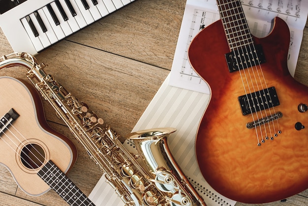 Composition of different musical instruments: synthesizer, electronic guitar, saxophone and ukulele lying, sheets with music notes lying on and wooden floor. Musical instruments. Music equipment