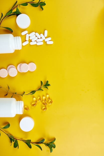 Composition of pills, fish oil, vitamins with green leavws on yellow surface