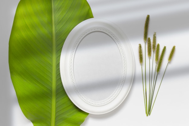 Composition with round oval white blank wooden frame laying on green leaf with field plants