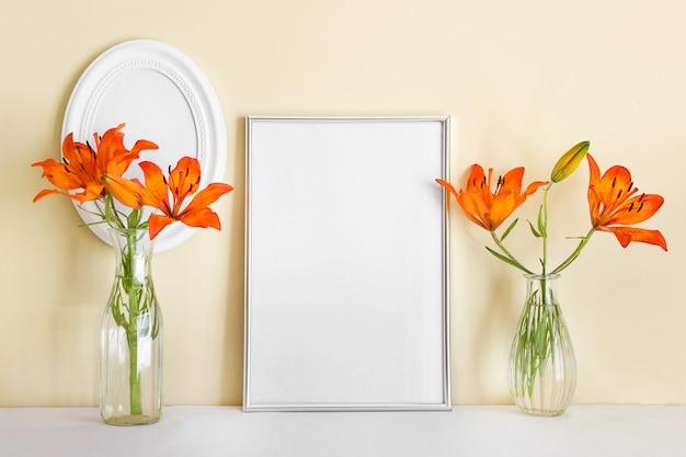 Composition with standing blank empty silver A4 frame and and orange lilies in glass vases.