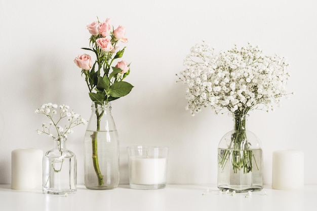 Composition with white and pink flowers in glass bottles and candles on table wall background.