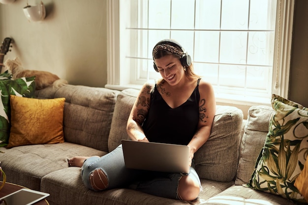 Computer headphones and woman on sofa and music happy work from home for mental health in apartment Young person relax on couch and listening to audio technology electronics and working on laptop