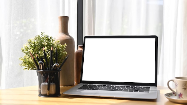 Computer laptop with white empty screen pencil holder and coffee cup on wooden table