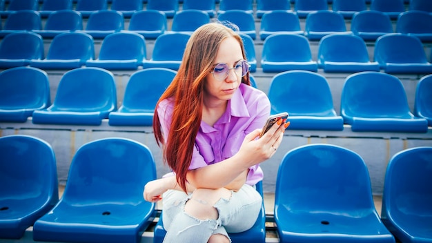 Concentrated adult female with long brown hair in blouse and eyeglasses sitting on seat of blue tribune and messaging on smartphone