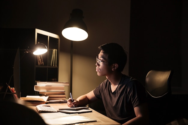 Concentrated asian young man in glasses using computer for studying and writing in notepad in the evening at home