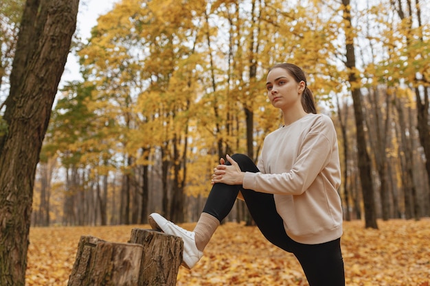 Concentrated athletic female runner stretching legs and leaning on tree while training in fall