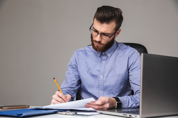 Concentrated bearded elegant man in eyeglasses writing something while sitting by the table in office