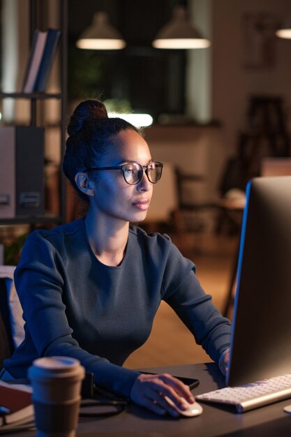 Concentrated beautiful multiethnic businesswoman in glasses sitting at desk in dark office and working with online documents on computer