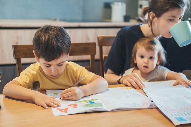 Concentrated boy doing his homework sitting at the table with mother who helping him concept of fami