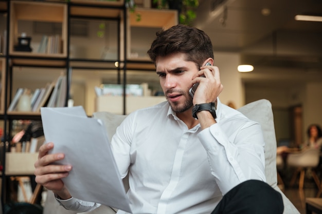 Concentrated businessman dressed in white shirt sitting in cafe and talking by phone while looking at documents.