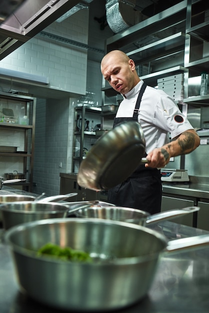 concentrated chef  with several tattoos on his arms holding a frying pan in a restaurant kitchen