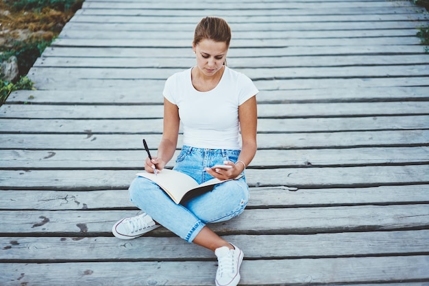 Concentrated clever hipster girl sitting outdoors and using mobile phone for blogging young