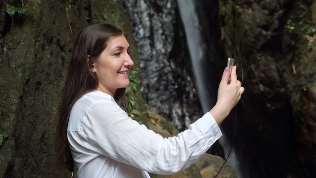 Concentrated dark haired girl blogger wearing headphones holds smartphone in hand and conducts video call against tropical waterfall close view