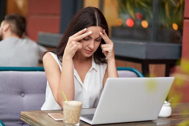 Concentrated female journalist tries to focuse as creats new article, sits in front of modern laptop computer at cafeteria