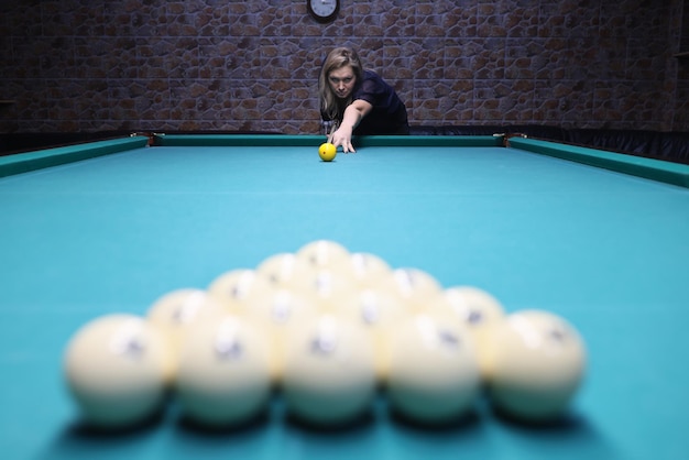 Concentrated female player directs ball to white pyramid made on blue pool table in dark room