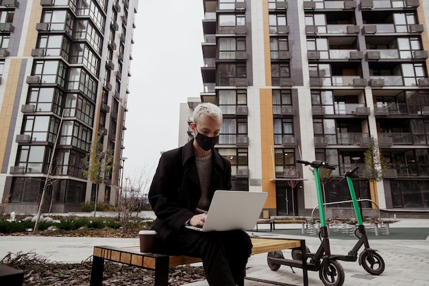 Concentrated freelancer man in black face mask working outside in park and using his modern laptop. He sits on bench and writes a program. Two electric scooters near the bench. Apartment blocks.