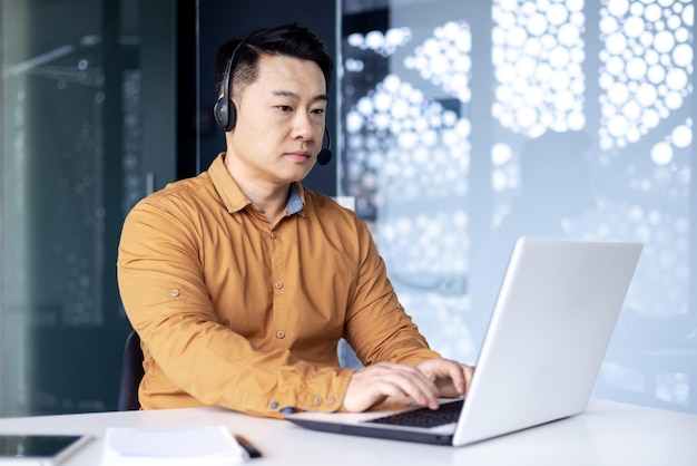 Concentrated korean office worker in headset sitting at desk in front of laptop in modern office