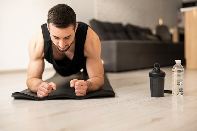 Photo concentrated man doing plank position while exercising on black yoga mat. water bottles lying near. smart modern interior on backgroung. man in black sportswear exercising at home.