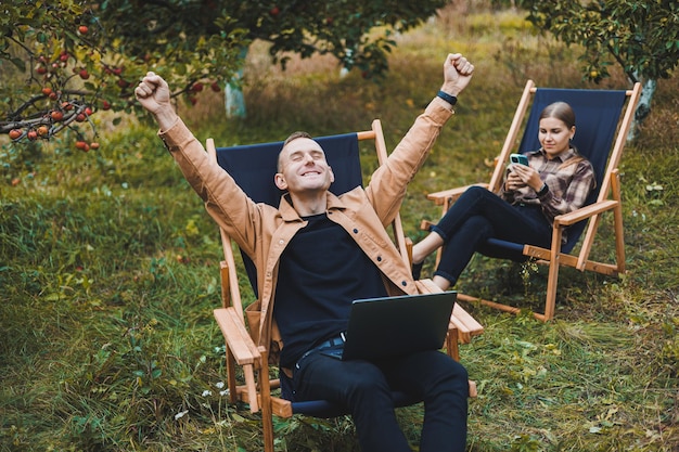 Concentrated man in the garden working on a laptop while sitting on a chair among the trees a man