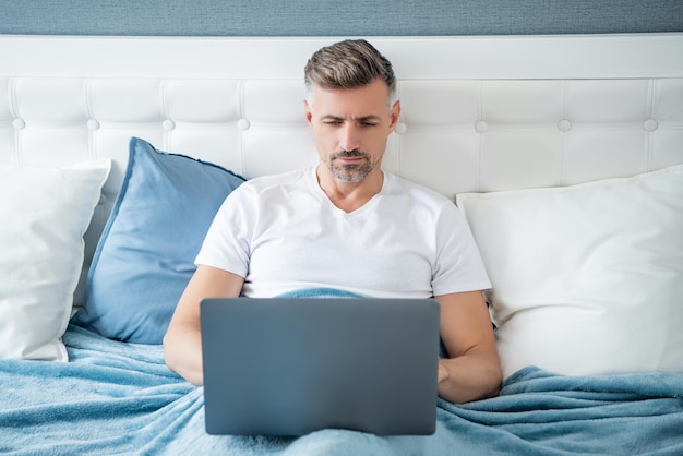 Concentrated mature man working on laptop in bed