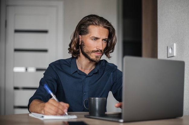 Concentrated middle aged man working on laptop and writing in notepad