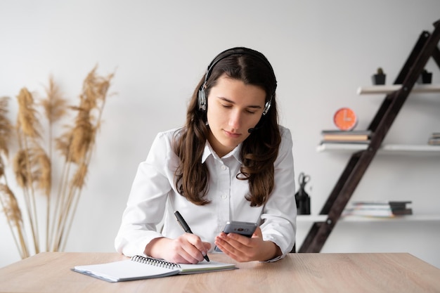 Concentrated millennial girl in a wireless headset does her homework while sitting at her desk at home studying online distance from the phoneCollege student listens to audio recording in a notebook