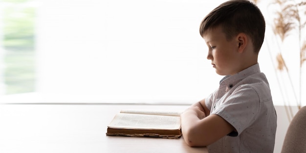 Concentrated motivated boy student sitting by the window at his desk reading a textbook in a lesson at school or home class Schoolboy reads a book near the place for the text school kids