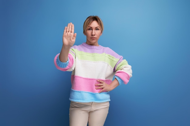 Concentrated pleasant blond young woman in a striped sweater shows stop with her hand on a blue