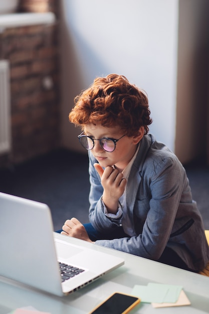 Concentrated. Red-haired boy in sitting at the laptop and looking concentrated