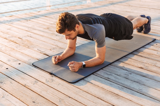 Concentrated sportsman in tracksuit lying on mat, and doing plank while working out on wooden pier at seaside in morning