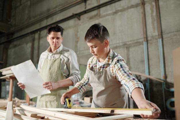 Photo concentrated teenage son in apron using tape measure while checking size of wooden plank, he working with father in carpentry shop