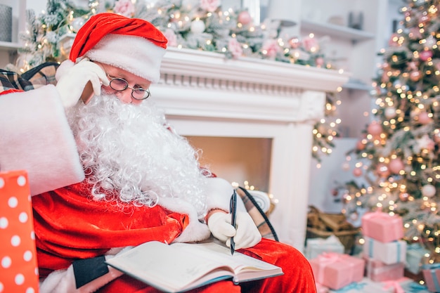 Concentrated and thoughtful Santa Claus sits at fireplace and Christmas tree writing in a notebook