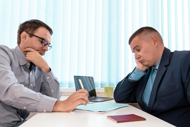 Concentrated two businessmen sitting at table involved in serious negotiations or contract discussion in blue office Focused young male ceo executive manager communicating with partner or client